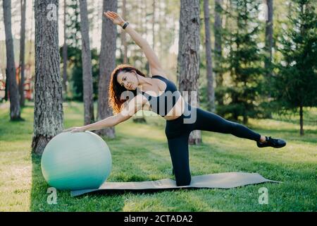 Colpo all'aperto di bruna attiva donna in abbigliamento sportivo posa su tappetino yoga, fa esercizi di stretching con palla ginnica, posa in foresta o parco su verde Foto Stock