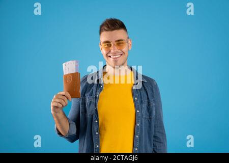 Sorridente giovane turista maschile con passaporto e biglietti su sfondo blu studio. Concetto di viaggio aereo Foto Stock