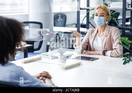 Donna afroamericana parla alla donna d'affari in maschera protettiva attraverso la partizione in vetro all'interno dell'ufficio Foto Stock