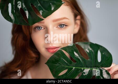 Giovane donna dai capelli rossi che posa tenendo le foglie tropicali sopra sfondo grigio Foto Stock