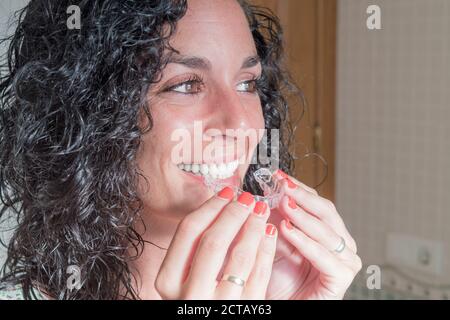 Primo piano di giovane donna di bruna con capelli ricci che mette sul suo allineatore invisibile in silicone. Correzione dentale. Ortodonzia mobile Foto Stock