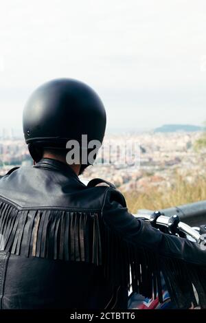 vista posteriore di un biker irriconoscibile con casco e giacca in pelle che guarda la città da un punto di vista, concetto di libertà e stile di vita ribelle, Foto Stock