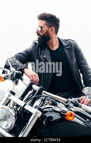 foto verticale di un uomo con giacca in pelle e occhiali da sole che si appoggiano alla sua moto, concetto di libertà e stile di vita biker, spazio copia per t Foto Stock