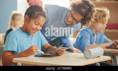 Scuola elementare computer Science Class: Smart Girl utilizza il computer Tablet digitale, un insegnante amichevole la aiuta spiegando lezione e Assignment Foto Stock