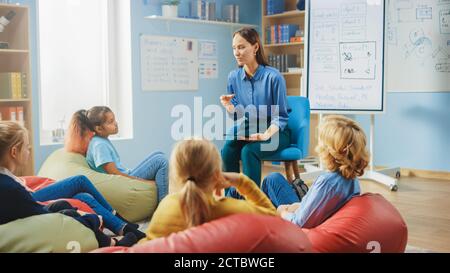 Classe di creatività della scuola elementare: I bambini seduti sui sacchi dei fagioli mentre l'insegnante gentile spiega la lezione mentre usa il computer del tablet digitale. Intelligente Foto Stock