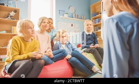 Classe di creatività della scuola elementare: I bambini seduti sul tappeto mentre l'insegnante premuroso spiega la lezione. Bambini intelligenti imparare in amichevole moderno Foto Stock