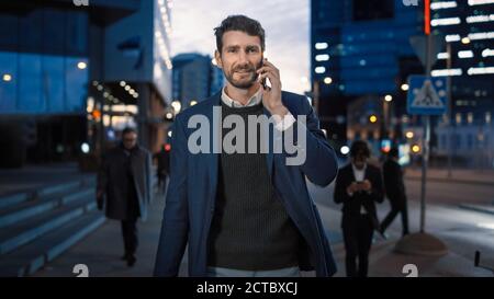 Uomo d'affari alto in un vestito sta parlando su un telefono mentre camminando nella città. Sta avendo una conversazione piacevole. Persone in ufficio a piedi. È sera Foto Stock