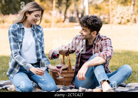 uomo che tiene una bottiglia e versa vino rosso in un bicchiere vicino donna seduta su una coperta di plaid nel parco Foto Stock