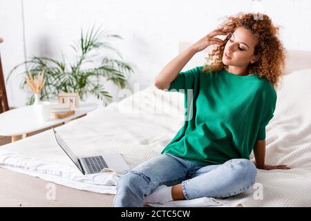 Giovane donna che tocca i capelli vicino al computer portatile sul letto Foto Stock