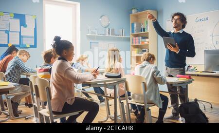 Insegnante entusiasta che dà una lezione in scuola elementare a classe piena di bambini diversi. Gruppo di bambini multietnici intelligenti che imparano nuove cose Foto Stock