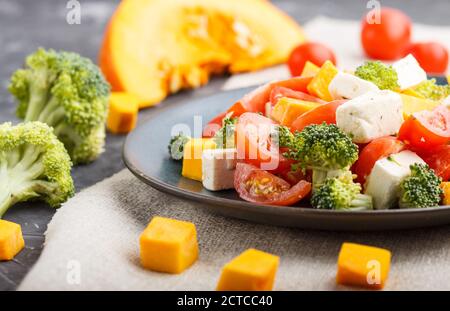 Insalata vegetariana con broccoli, pomodori, formaggio feta e zucca su una piastra di ceramica blu su fondo nero di cemento e tessuto di lino, vista laterale Foto Stock