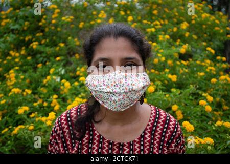 Bella donna con maschera floreale, orecchini e top stand designer prima di fiori gialli. Protezione dalla corona in modo elegante. Foto Stock