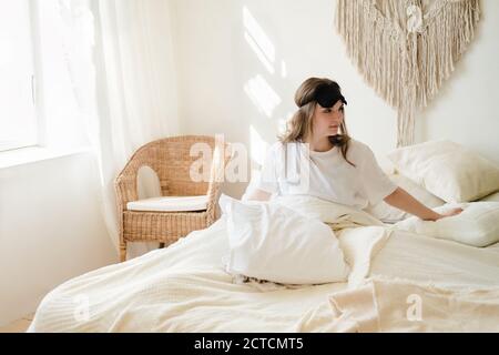 Giovane bella donna in comodo letto e maschera per dormire la mattina in casa accogliente. Autoisolamento, soggiorno a casa durante la quarantena, pandemia Foto Stock