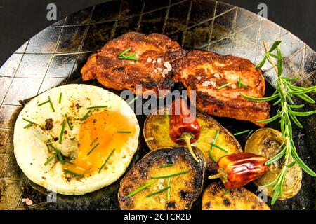 Closeup di costolette di maiale fritte nella padella sul fornello decorato con rosmarino. Collo di maiale con uova e patate. Carne fritta. Foto Stock