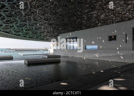 Una foto dall'interno del Louvre Abu Dhabi al bordo delle acque, che presenta il suo tetto ad alta tecnologia che mostra la luce che filtra attraverso e intorno sull'acqua, la sua facciata bianca che mostra le pareti. Aperto a novembre 2017. Foto Stock