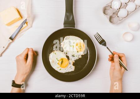 Vista dall'alto delle mani femminili che tengono la forchetta sulla padella con uova fritte tra gli ingredienti da cucina per una sana colazione su un tavolo bianco. Foto Stock