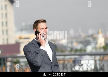 Imprenditore sorridenti utilizza lo smartphone comunicazione, skyline background. Uomo in tuta imprenditore assume i vantaggi delle moderne tecnologie mobili. Business il concetto di chiamata. L'uomo parla cellulare all'aperto. Foto Stock