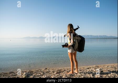Giovane fotografo femminile con treppiede sulla spalla, fotocamera in mano e zaino fotografico sulla schiena in piedi su bella spiaggia di ghiaia vicino Foto Stock