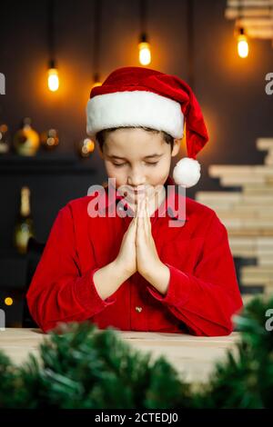 Ragazzo carino che prega e sogna un regalo per Natale Nel cappello rosso di Santa Foto Stock