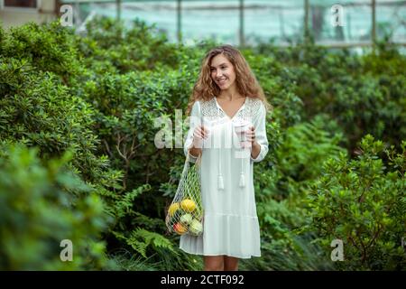 Giovane donna in abito bianco con sacchetti di verdure e. una tazza in mani Foto Stock