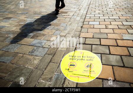 Il volto che cambia della strada alta. Una persona getta un'ombra a piedi davanti a un segnale di informazione di allontanamento sociale dipinto sul marciapiede nel centro della città di Leeds, a circa sei mesi dalla sera di marzo 23, quando il primo ministro Boris Johnson ha annunciato restrizioni a livello nazionale. Foto Stock