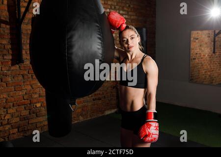 Indossa una robusta bruna donna con top sportivo e pantaloncini corti una borsa da punzonatura con guanti da boxe in palestra Foto Stock