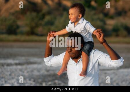 Bambino ragazzo seduto sul collo del padre e sorridente. Bel giovane padre afro-americano e suo figlio carino. Padre e figlio trascorrono il tempo insieme Foto Stock