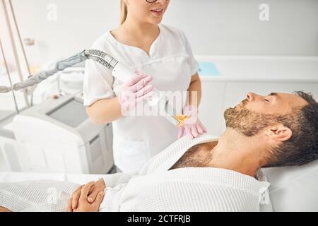 Affascinante estetista femminile che tiene il dispositivo laser di rimozione dei capelli e sorridente mentre il giovane sopportato si stese sul letto da giorno Foto Stock