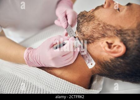 Primo piano di donne estetiste mani in guanti sterili mettere righello sul collo maschio e linea retta con bianco matita Foto Stock