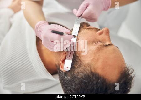 Primo piano di mani estetista donna in guanti sterili posizionamento righello su guancia maschio e disegno in linea retta con bianco matita Foto Stock