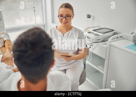 Bella donna cosmetologa che guarda paziente maschile e sorridente mentre tenendo gli appunti Foto Stock