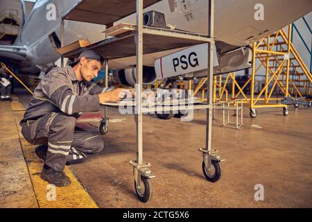 Primo piano ritratto in vista laterale di giovane meccanico tecnico seduto hunker giù mentre usando l'attrezzatura speciale nell'hangar dell'aviazione Foto Stock