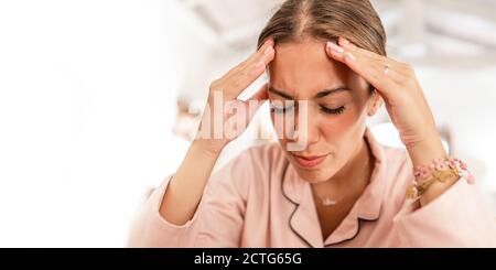 Una giovane donna caucasica con pajama rosa tiene la testa nelle sue mani e grimaces nel dolore dall'emicrania mal di testa Foto Stock