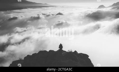 Una scala di grigi di un uomo solitario in piedi sul bordo di una montagna di fronte alle nuvole durante la luce del giorno Foto Stock