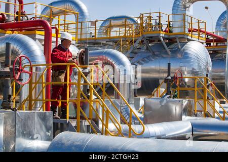 Regione di Aktobe/Kazakhstan - Maggio 04 2012: Stabilimento di raffineria di petrolio. Tubi metallici lucidi, tubi, scambiatori di calore, addetti alla raffineria o ingegneri in rosso usura da lavoro WIT Foto Stock