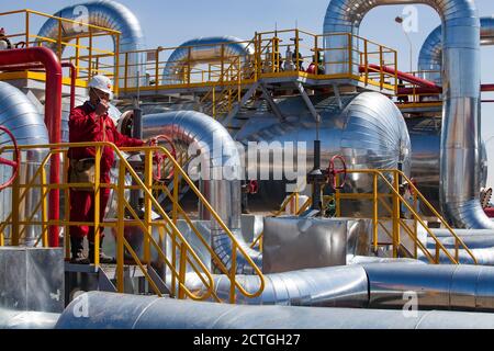 Regione di Aktobe/Kazakhstan - Maggio 04 2012: Stabilimento di raffineria di petrolio. Tubi metallici lucidi, tubi, scambiatori di calore, addetti alla raffineria o ingegneri in rosso usura da lavoro WIT Foto Stock