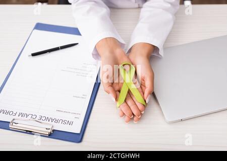 vista ritagliata dello psicologo che tiene nastro di consapevolezza verde vicino forma di reclamo di assicurazione e laptop, concetto di salute mentale Foto Stock