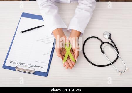 vista dall'alto dello psicologo che tiene un nastro di consapevolezza verde vicino alla forma di reclamo assicurativo e stetoscopio, concetto di salute mentale Foto Stock