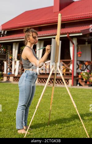 Giovane bella donna pittura immagine su cavalletto in prestito in campagna, hobby Foto Stock