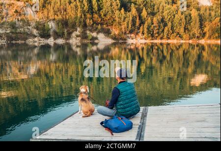 Uomo come proprietario di cane e il suo amico cane beagle sono seduti sul molo di legno sul lago di montagna e godendo il paesaggio durante la loro passeggiata in th Foto Stock