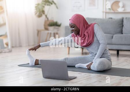Formazione a casa. Donna musulmana nera sportiva che si allunga davanti al computer portatile Foto Stock