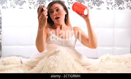 Bella giovane donna si sveglia a letto con una tazza di caffè e una sveglia. La donna ha dormito troppo ed è in ritardo, reagendo con orrore al momento. Foto Stock