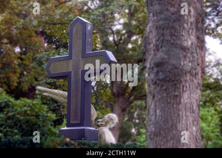 Croce cristiana di pietra di fronte ad un albero e un angelo scultura, fuoco selettivo sulla croce Foto Stock