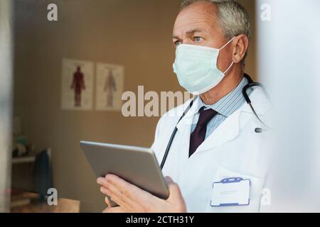 Medico anziano in piedi indossando maschera utilizzando tablet digitale in ospedale Foto Stock