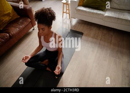 Vista ad alto angolo della giovane donna africana in posizione di loto pratica di meditazione e respirazione esercizio a casa Foto Stock