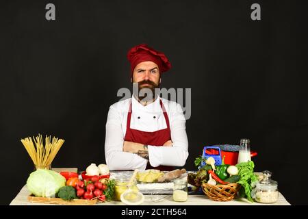 Chef con pasta, verdure e pasta sul tavolo. Concetto di cibo fatto in casa. L'uomo con la barba si siede dal piano di lavoro su sfondo nero. Cuocete con il viso stretto in uniforme di borgogna vicino agli ingredienti. Foto Stock