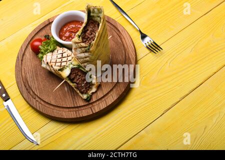 Pane piatto con ripieno di maiale o pollo e salsa di peperoncino rosso. Concetto di fast food. Burrito o tortilla serviti in ristorante o caffè. Carne tritata avvolta con pane di lavasino su sfondo di legno giallo. Foto Stock