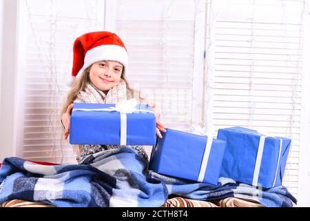 Santas helper in cappello rosso coperto di coperta blu contiene scatole regalo blu. Ragazza con viso generoso si siede su sfondo chiaro finestra. Il bambino si prepara alle vacanze invernali. Natale e concetto di infanzia Foto Stock