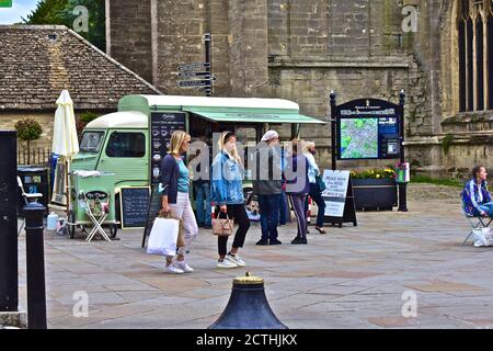 Le persone che passeggiano attraverso il Cirencester Market Place, con un vecchio van Citroen HY francese in uso come una stalla di caffè. Chiesa di San Giovanni Battista alle spalle. Foto Stock