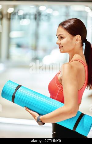 Vista laterale di una giovane donna attraente in abbigliamento sportivo con tappetino yoga e guardando lontano mentre si trova in studio prima di lezione di yoga o di allenamento fitness. Sport, benessere e stile di vita sano Foto Stock